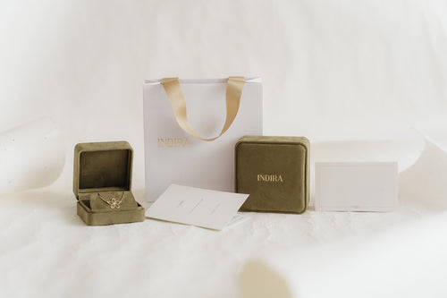 Set per confezione regalo Indira: shopper bianca con manici in nastro dorato, due cofanetti portagioielli in velluto color oliva (uno aperto che mostra un pendente a forma di fiore in oro) e cartoncini brandizzati su un morbido sfondo bianco da studio.