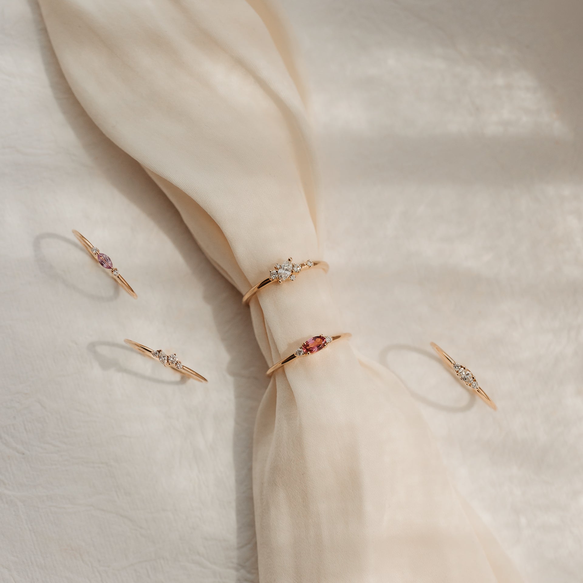 Cinque delicati anelli in oro giallo 14k con diamanti e gemme rosa, disposti su un tessuto di seta color crema annodato alla luce solare soffusa.