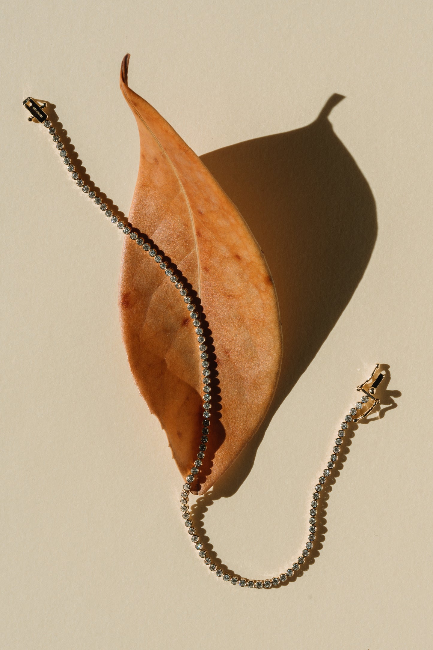 Un bracciale tennis in oro con diamanti poggiato su una foglia autunnale su sfondo beige.