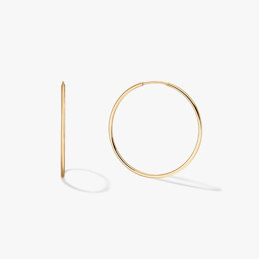 Coppia di grandi orecchini a cerchio sottili in oro giallo 14K su sfondo bianco, uno visto frontalmente e uno di profilo con ombre morbide.