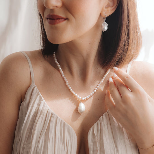 Primo piano di una modella che indossa un elegante orecchino pendente con perla barocca in argento 925 rodiato, con un piccolo dettaglio in zirconia, e una collana di perle coordinata; la mano è vicino alla clavicola, in una morbida luce naturale.