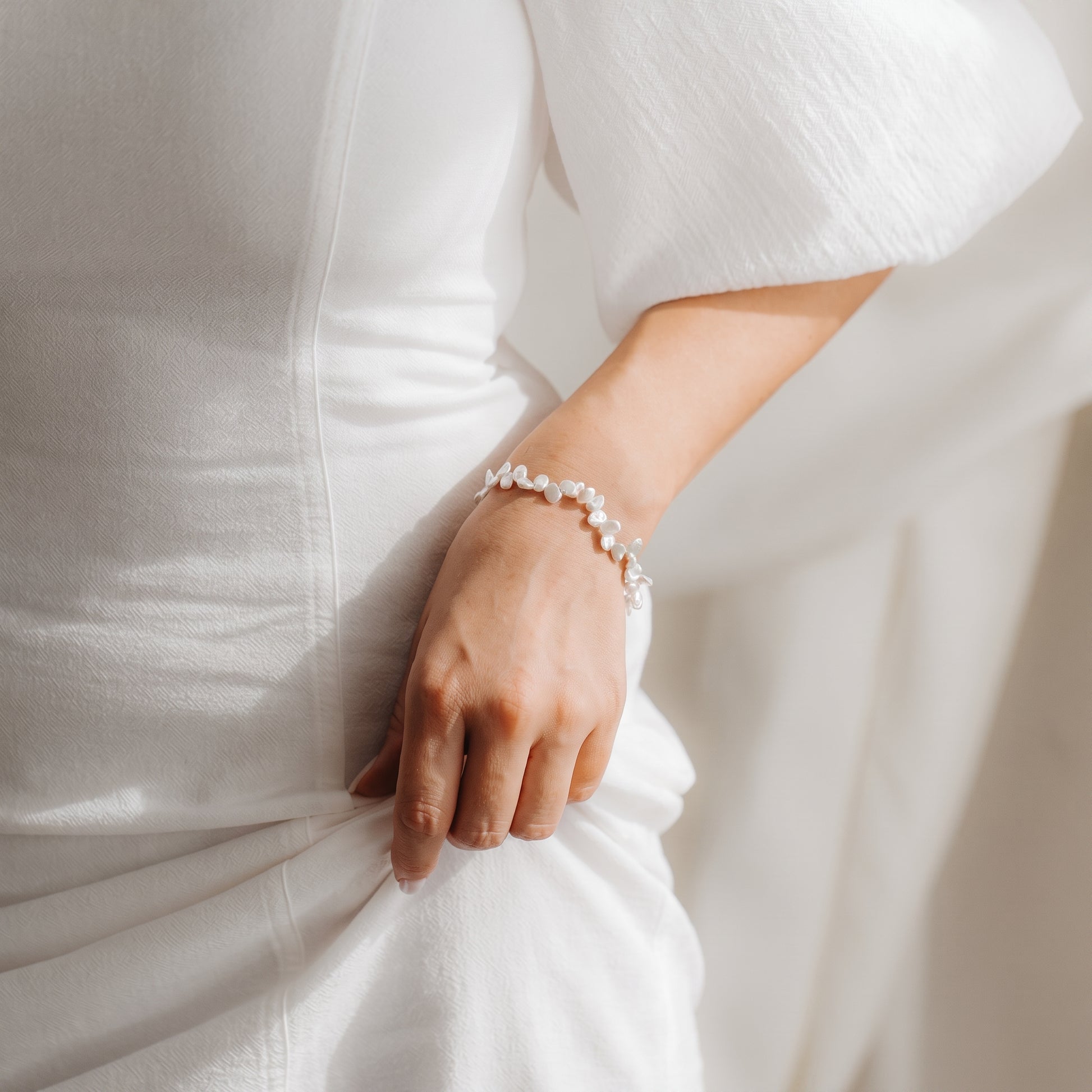 Primo piano di un polso con un bracciale con perle Keshi in argento sterling placcato oro, abbinato a un abito bianco in una soffusa luce naturale.
