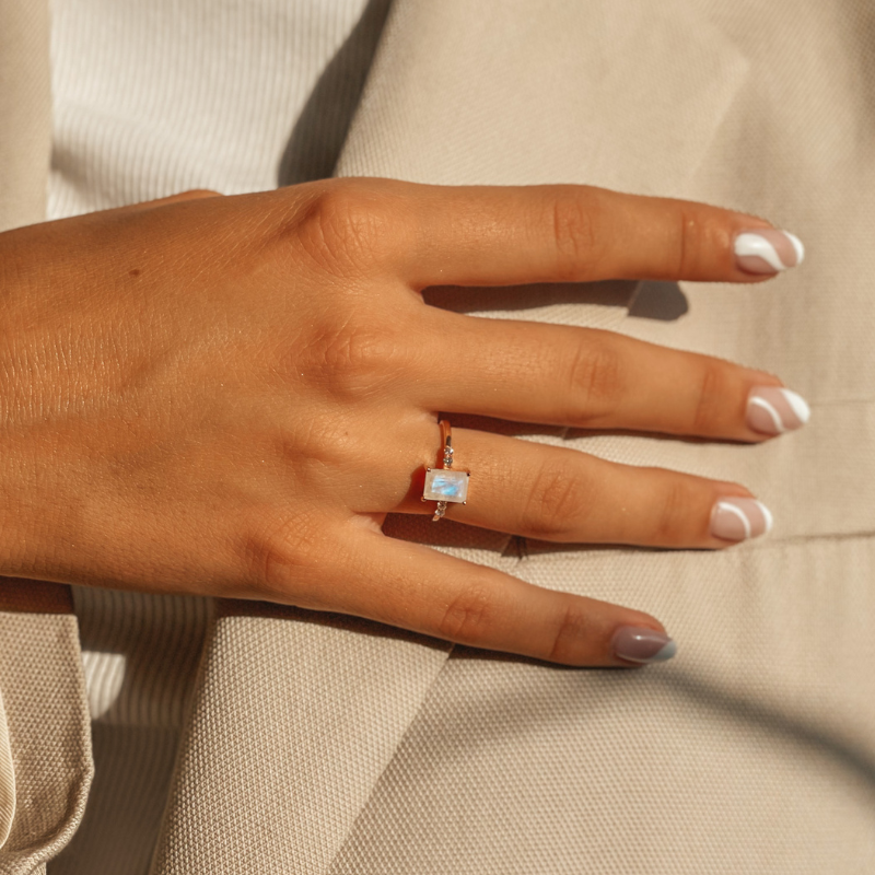 Primo piano di una mano che indossa un anello in oro rosa con pietra di luna su uno sfondo di tessuto beige.