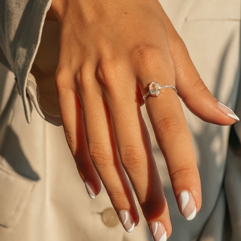 Primo piano di una mano che indossa un anello in argento con pietra lunare su uno sfondo beige.