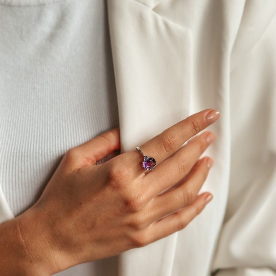 Primo piano di una mano che indossa un anello d'argento con gemme di ametista e tanzanite, appoggiata su un indumento bianco.